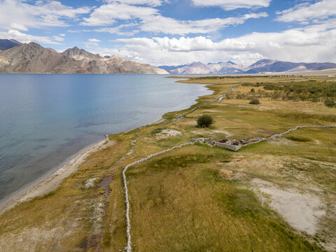 Aerial view of Pangong Lake with its deep blue waters, surrounding arid mountains, and a stone wall boundary along the grassy shoreline in Merak, Ladakh, India.
