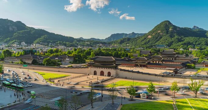 Busy street traffic streaks past Gwanghwamun Gate and Gyeongbokgung Palace in Seoul, South Korea beneath a blue sky with mountains rising behind the historic complex - timelapse high angle