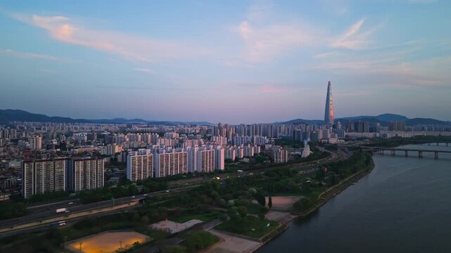 Aerial parallax shot over Gwangnaru Han River Park at dusk with expressway traffic, floodlit field and Lotte Tower glowing pink on horizon