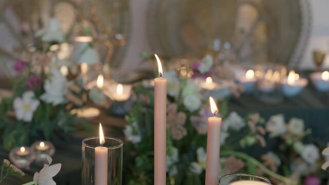 Lit taper candles and glass hurricanes among white flowers and greenery on a banquet table