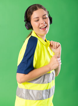 Girl worker wearing high visibility polo listens to music using headphones and dancing. Female person on green background or isolated human figure