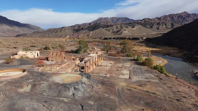 Drone footage captures the abandoned sulfate de aluminio plant ruins in Calingasta, Argentina, surrounded by rugged Andean mountains, a clear sky, river, and arid landscape.