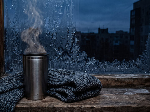thermal mug with hot steam and a knitted scarf standing on a windowsill in front of a frosty window