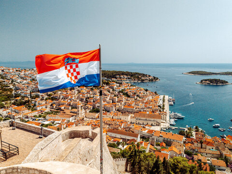 Aerial view of the Croatian flag waving over the historic Hvar Port, featuring terracotta rooftops, the Adriatic Sea, and boats in the harbor in Hvar, Split-Dalmatia County, Croatia.