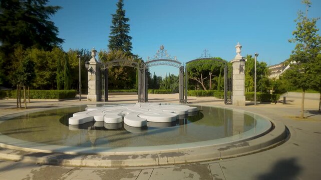 A contemporary white sculpture fountain sits in a reflecting pool before the ornate iron gates of El Retiro Park in Madrid. Ideal for travel documentaries and urban wellness content.