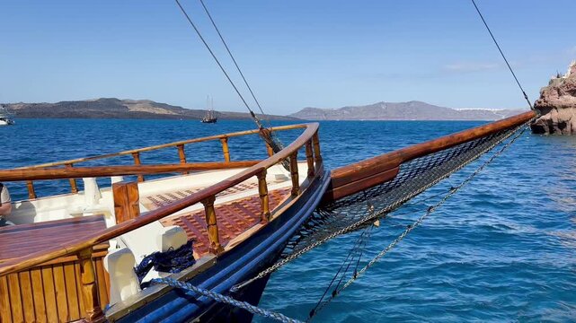 Wooden caldera cruiser sailing across deep blue Aegean waters between Nea Kameni and Thirasia in Santorini, Greece, with bowsprit, rigging and distant volcanic islands