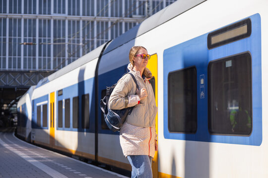 A female traveler stands at a train station wearing sunglasses and a backpack. She waits for her train in the bright sunlight.