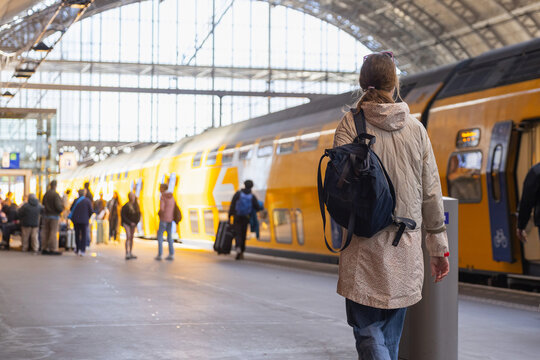 A female traveler walks towards a yellow train at a busy train station in daylight. People are waiting and preparing for travel.