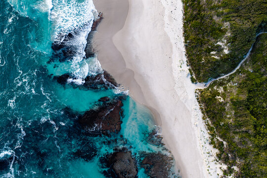 Aerial view of turquoise waves crashing against dark rocks and a white sandy beach bordered by lush vegetation in Nullaki, Western Australia, Australia.
