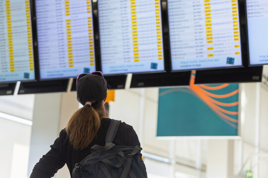 A female traveler looks at flight information screens in the airport terminal. She stands with a backpack facing the display boards.