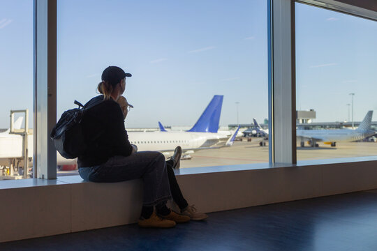 A female traveler sits comfortably with her child in an airport terminal while observing planes. She enjoys a drink and watches the busy activity outside.
