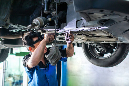 Selective focus to Professional mechanic wearing headlamp inspecting and repairing car chassis, suspension, and brake system underneath a lifted vehicle in garage.