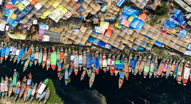 Aerial view of colorful wooden boats docked along the riverbank near a bustling market with vibrant tarpaulin shades in Tongi, Dhaka Division, Bangladesh.