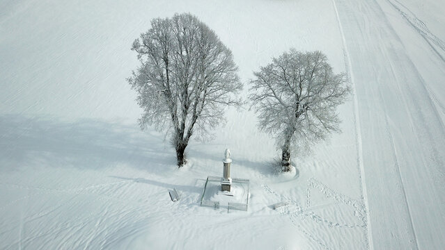 Aerial view of a monument surrounded by two frost-covered trees in a snow-covered field in Giron, Auvergne-Rhone-Alpes, France.