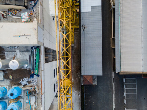 Aerial view of a construction site with a yellow tower crane between industrial buildings, blue water tanks, and materials on the ground in Purwokerto, Central Java, Indonesia.