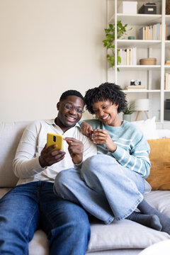 Happy couple using smartphone and smiling while relaxing on sofa at home