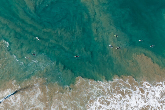 Aerial view of surfers waiting for waves in the turquoise ocean with sandy sediment patterns near the shore in Newport Beach, California, United States.