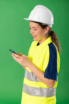 Polite young girl worker wearing high visibility polo and hard hat scrolling smartphone standing against green screen