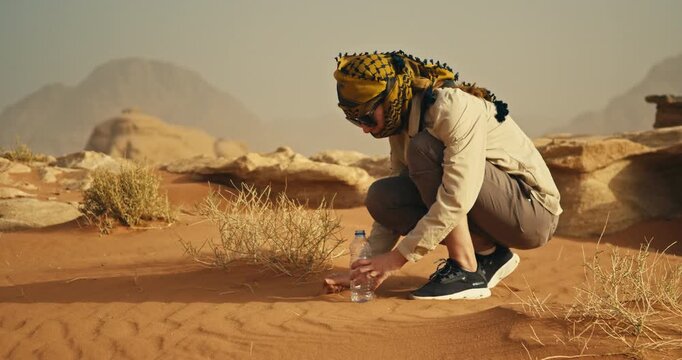 Person crouching in Wadi Rum, Jordan, carefully collecting golden desert sand in a clear bottle, surrounded by dry vegetation and weathered sandstone formations in warm sunlight.