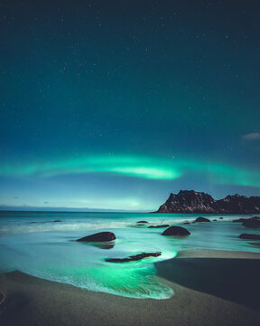 Aerial view of the stunning aurora borealis illuminating the night sky above the sandy beach and dark mountains of Lofoten, Nordland, Norway.