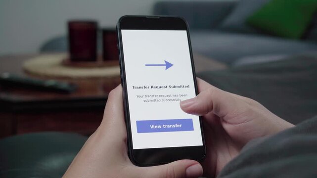 Person relaxes on a couch viewing a Transfer Request Submitted confirmation with a View transfer button on a smartphone.