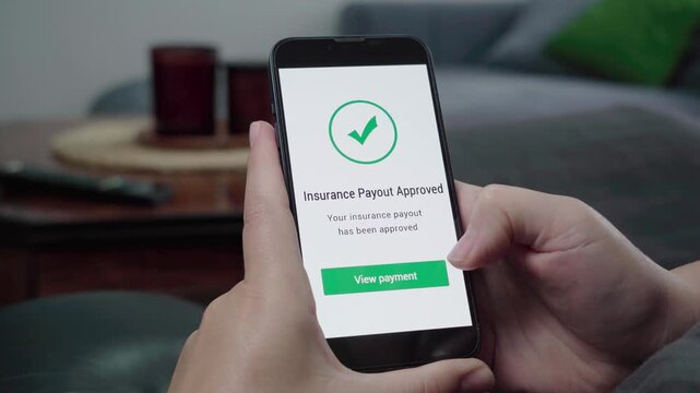 Hands hold a mobile device displaying an Insurance Payout Approved message with a green View payment call-to-action in a casual living room.