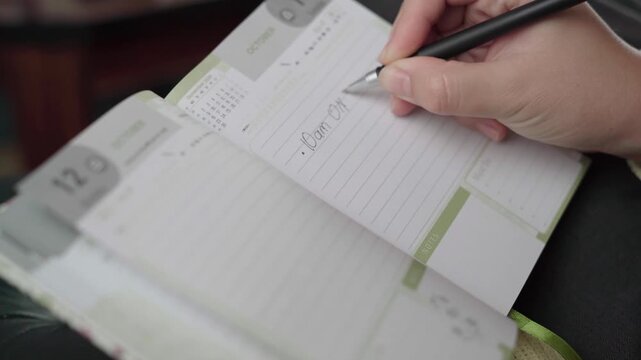Hand with pen writes an online meeting note into a dated daily organizer, showing remote work scheduling and time management.