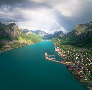 Aerial view of Gryllefjord nestled between steep mountains with a vibrant rainbow arching over the turquoise fjord waters and a small harbor in Gryllefjord, Troms, Norway.