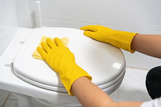 A person wearing yellow rubber gloves is meticulously cleaning a white toilet seat with a cloth.