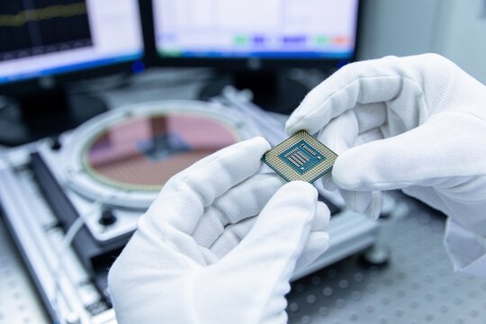 A close-up view of gloved hands holding a computer chip, with a semiconductor wafer in background.
