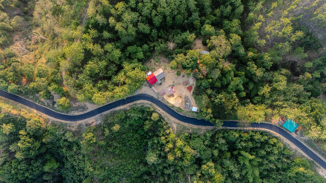 Bandarban, Bangladesh - 10 April 2024: Aerial view of a winding road cutting through a dense forest with a small building featuring a red roof nestled among the trees.