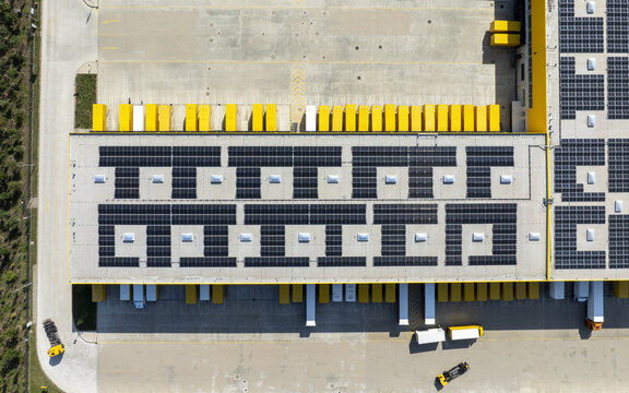 Aschheim, Germany - 20 July 2026: Aerial view of the DHL Paketzentrum Aschheim with solar panel arrays on the roof and yellow delivery trucks parked at the loading bays.