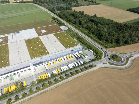 Aschheim, Bavaria, Germany - 20 July 2026: Aerial view of the DHL Paketzentrum Aschheim logistics facility with a green roof, loading docks, parked trucks, and surrounding agricultural fields.