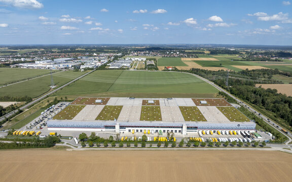 Aschheim, Germany - 20 July 2026: Aerial view of the DHL Paketzentrum Aschheim, a large logistics facility with a green roof, surrounded by fields and roads under a blue sky.