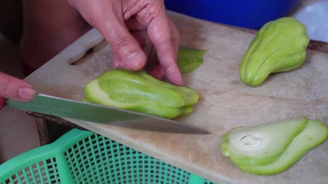 Close-up of hands slicing fresh green chayote on wooden cutting board with knife. Bright kitchen setting. Natural food prep concept for culinary or healthy eating themes.