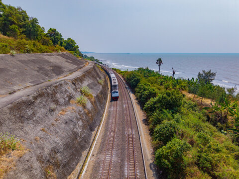 Aerial view of a train traveling along a coastal railway line with a concrete retaining wall and lush vegetation in Indonesia.