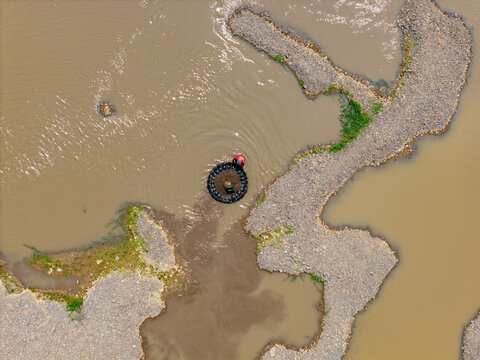 Aerial view of a fisherman in a small circular boat on a muddy river surrounded by gravel banks in Indonesia.