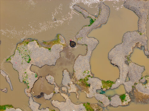 Aerial view of a person in a small circular boat navigating through muddy river waters surrounded by gravel and sand bars in Indonesia.