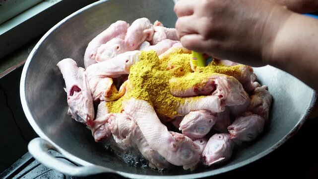 Close-up of hands adding vibrant yellow spice to raw chicken pieces in a metal wok. Natural lighting highlights textures and colors for culinary content, elegantly showcasing cooking.