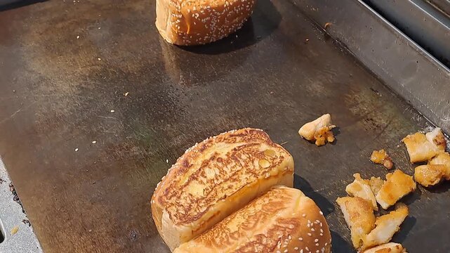 Close-up of a chef using tongs to toast buttered bread buns on a flat top grill alongside chicken pieces. Fast food preparation stock video footage.