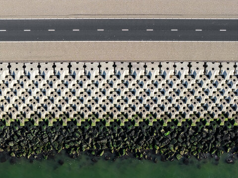 Aerial view of the Breezanddijk sea wall featuring interlocking concrete blocks, a paved road, and moss-covered rocks along the water in Breezanddijk, Friesland, Netherlands.