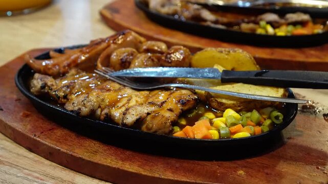 Grilled chicken steak with vegetables and potatoes served on a sizzling plate with cutlery ready to eat. Warm rustic wooden serving board adds texture. Perfect for restaurant or home cooking scenes.