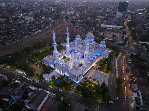 Aerial view of the illuminated Sheikh Zayed Grand Mosque Surakarta surrounded by urban streets, railway tracks, and city buildings in Kecamatan Banjarsari, Jawa Tengah, Indonesia.