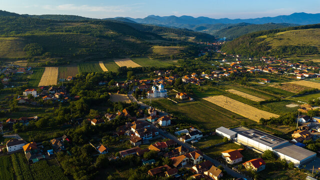 Aerial view of a village with a church featuring blue domes, surrounded by green hills, agricultural fields, and residential houses in Deva, Hunedoara, Romania.