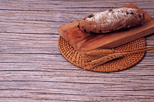 Fresh sourdough rests on wooden board, dusted with flour. Warm bread sits atop rustic cutting board. Wheat stalks lie beside it, woven mat below. Subtle shadow hints at soft, cozy kitchen light
