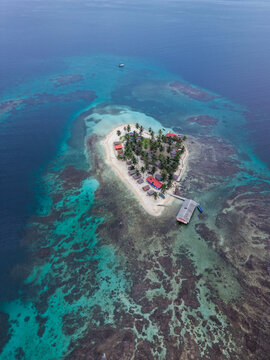 Aerial view of a small tropical island with palm trees, thatched-roof huts, and a wooden dock surrounded by turquoise coral reefs in Guna Yala Comarca, Panama.