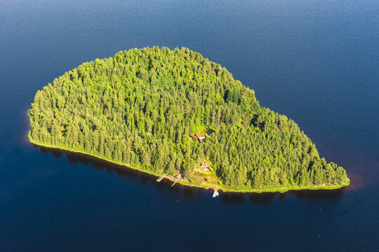 Aerial view of a lush green island covered in dense pine forest with small wooden cabins and a small dock, surrounded by the deep blue waters of Lake Siljan in Mora, Dalarna County, Sweden.