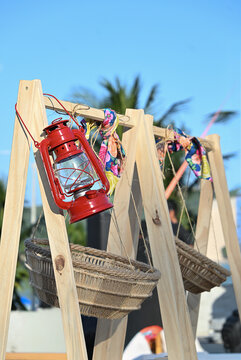 vintage red kerosene lantern hanging on rustic wooden display stand, outdoor party decor detail with suspended wicker baskets and palm tree background
