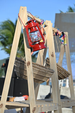 vintage red kerosene lantern hanging on rustic wooden display stand, outdoor party decor detail with suspended wicker baskets and palm tree background