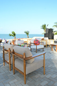 outdoor beach lounge deck with wooden armchairs and colorful patterned cushions, seaside event setup on paved stone terrace with ocean view and palm trees